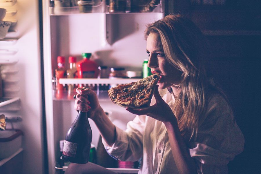 Frau steht im Dunkeln vor Kühlschrank und isst Pizza und trinkt Wein 
