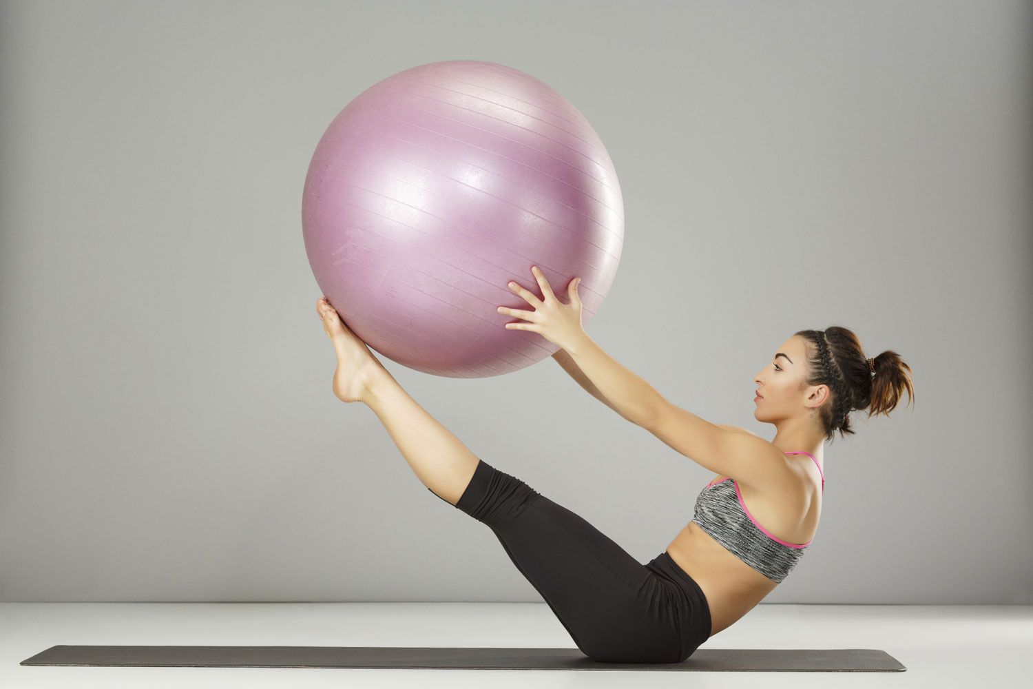 Gymnastikball-Übungen für den Bauch: das Klappmesser Athletische Frau trainiert mit Gymnastikball vor grauem Hintergrund.