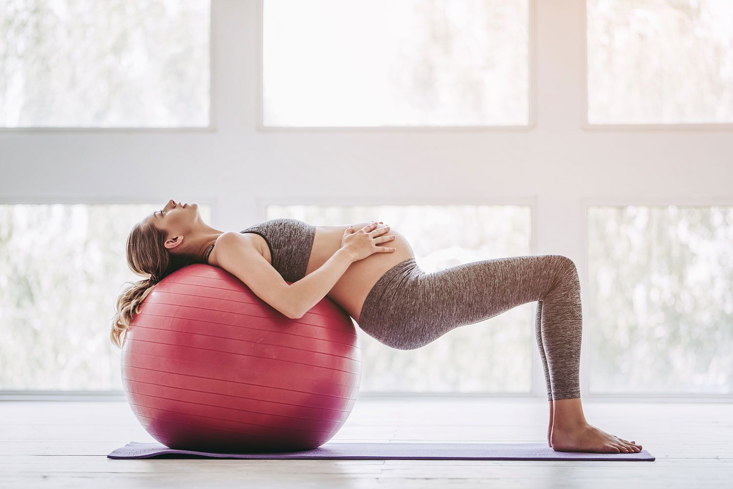 Schwangere Frau in Sportkleidung lehnt sich auf einen roten Gymnastikball, um den Rücken zu entlasten.