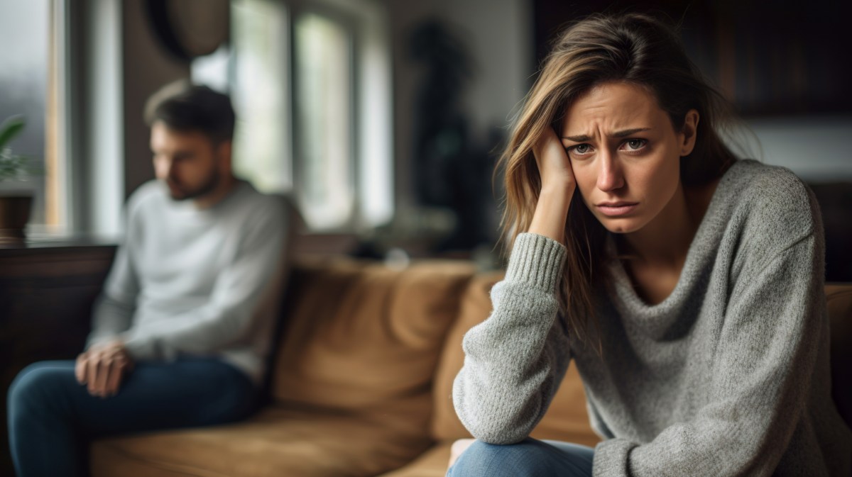 Frau auf einer Couch, die traurig und besorgt nach vorne schaut, während im Hintergrund ein Mann verschwommen auf der anderen Seite der Couch sitzt