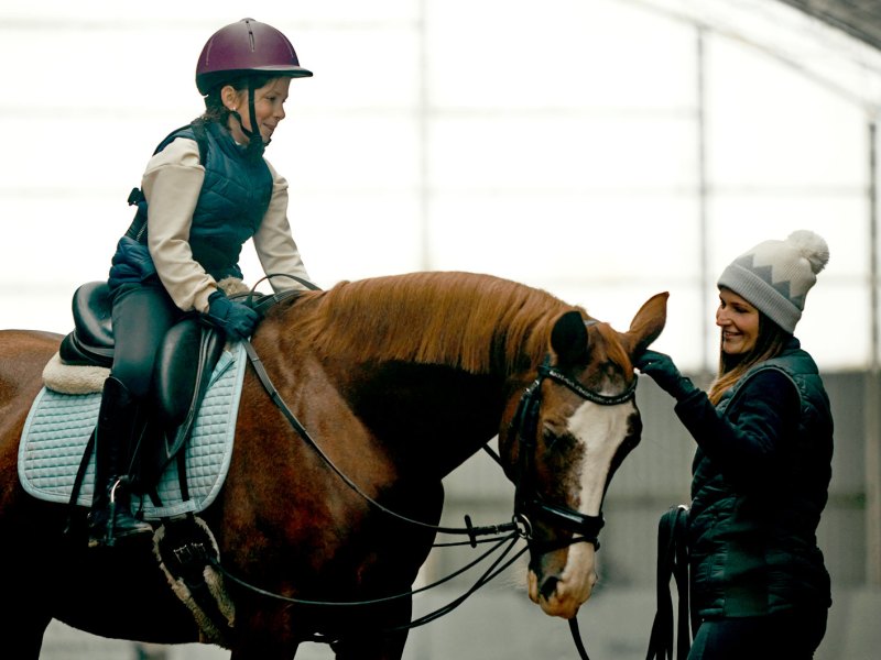 Mädchen auf einem Pferd in einer Reithalle. Die Reitlehrerin steht neben dem Pferd und streichelt es am Kopf.