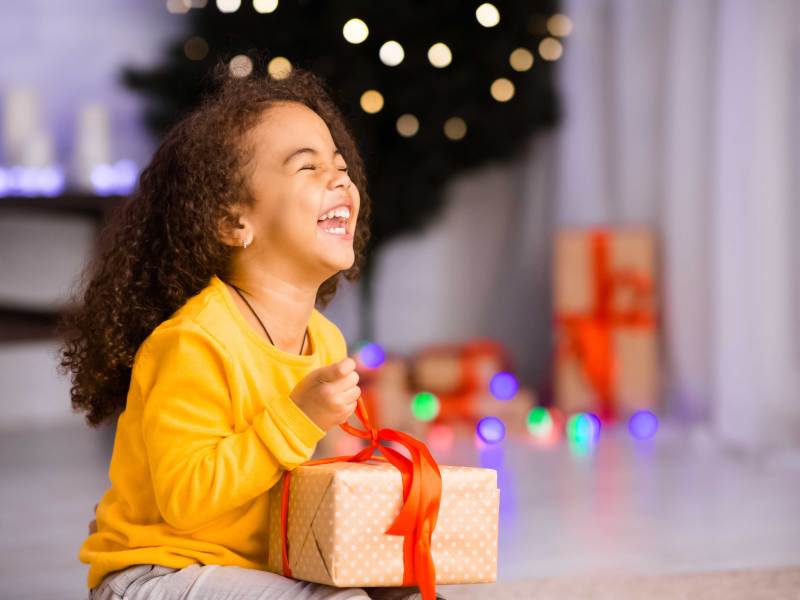 Kleines Mädchen sitzt vorm Weihnachtsbaum und packt sehr aufgeregt ein Geschenk aus.