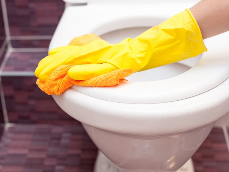 Person schrubbt eine Klobrille mit einem gelben Schwamm und trägt dabei gelbe Gummihandschuhe