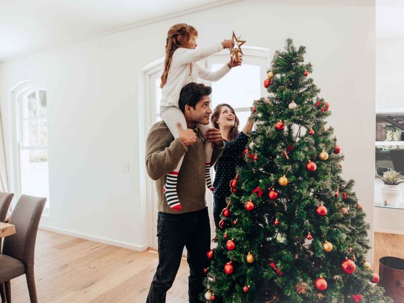 Vater, Mutter und Tochter schmücken gemeinsam den Weihnachtsbaum in ihrem Wohnzimmer. Der Vater trägt seine Tochter auf den Schultern, damit sie den Weihnachtsstern an der Baumspitze befestigen kann.
