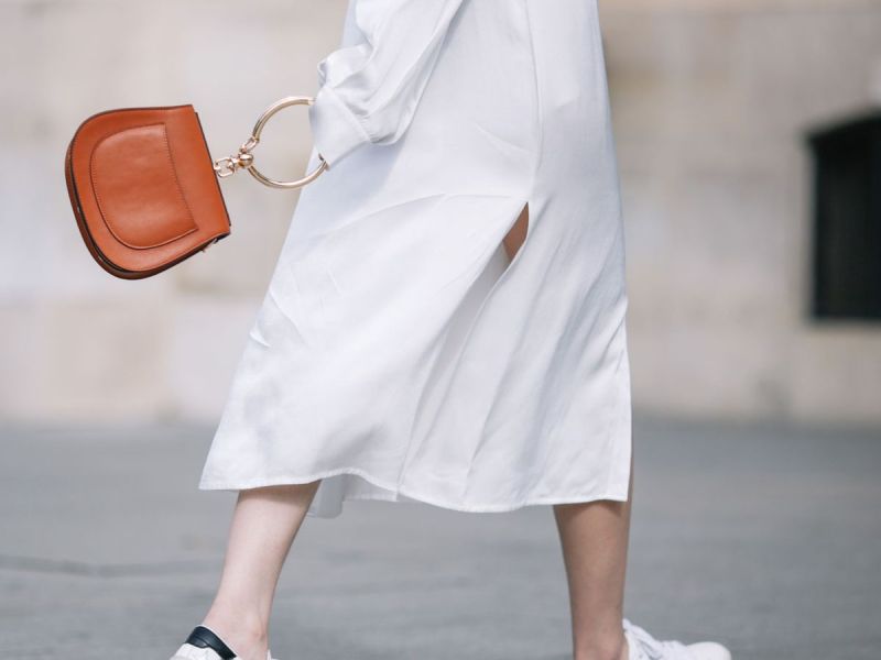 Frau im weißen Kleid schlendert mit einer Tasche und Sneaker in der Stadt
