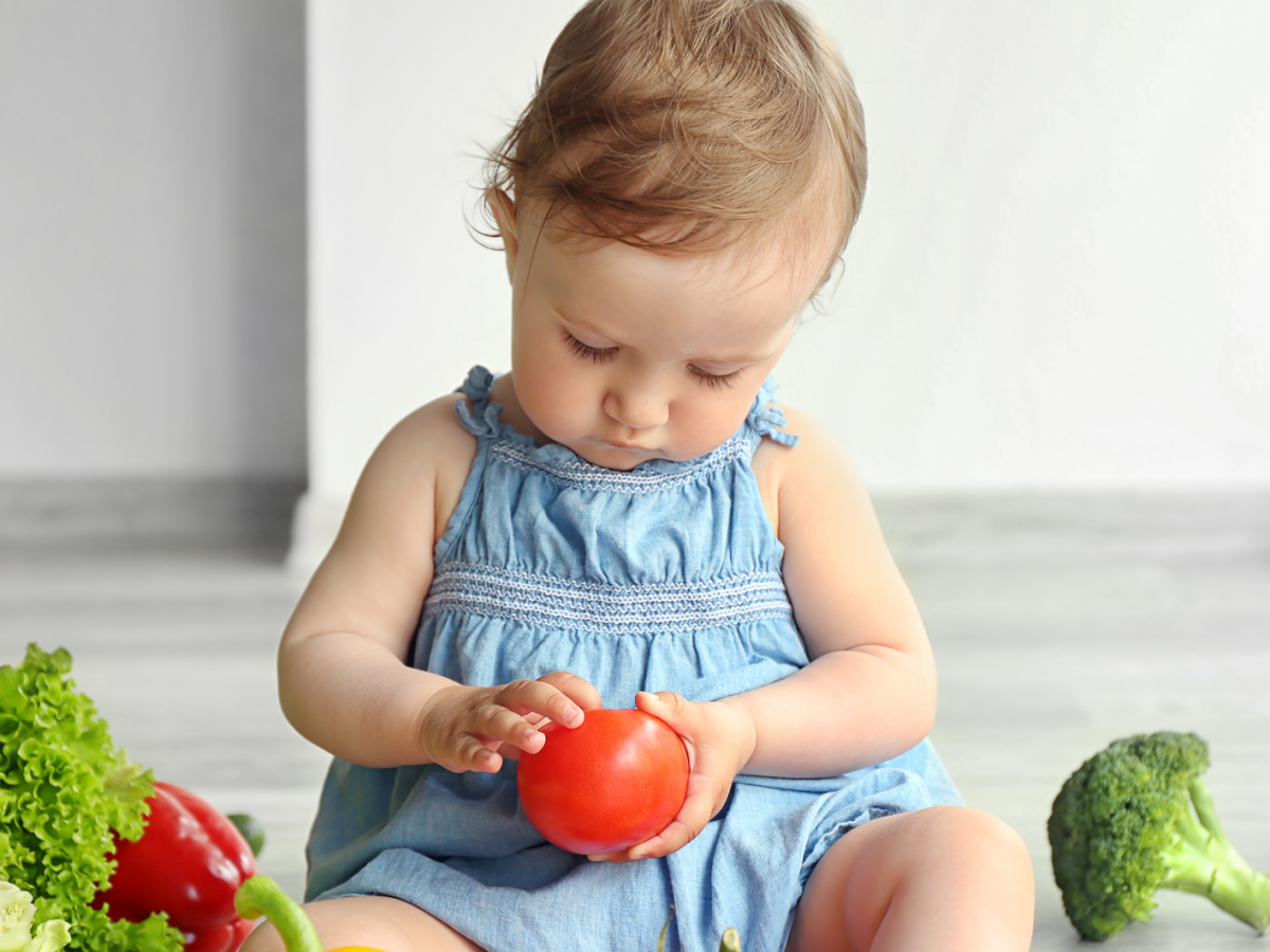 Kleinkind sitzt auf dem Boden und hat Tomaten und anderes Gemüse vor sich liegen