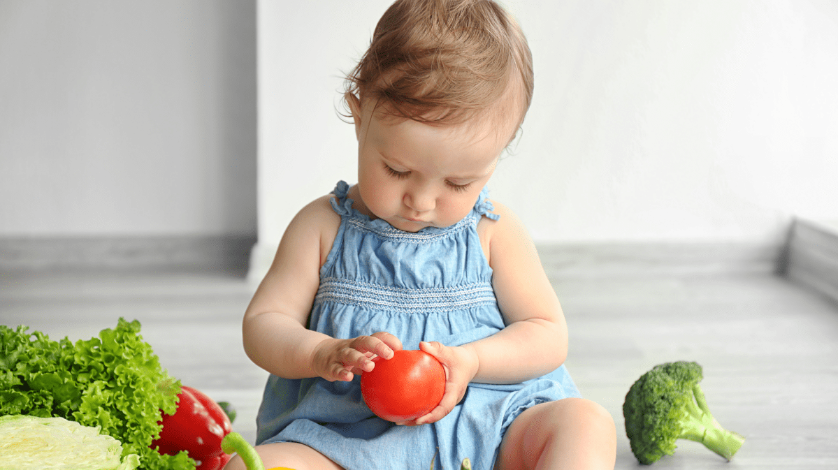Kleinkind sitzt auf dem Boden und hat Tomaten und anderes Gemüse vor sich liegen