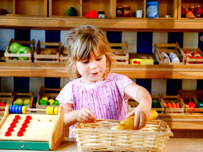 Kleines Mädchen steht in ihrer Holzspielküche und legt ein Spielzeugbaguette in einen Korb.