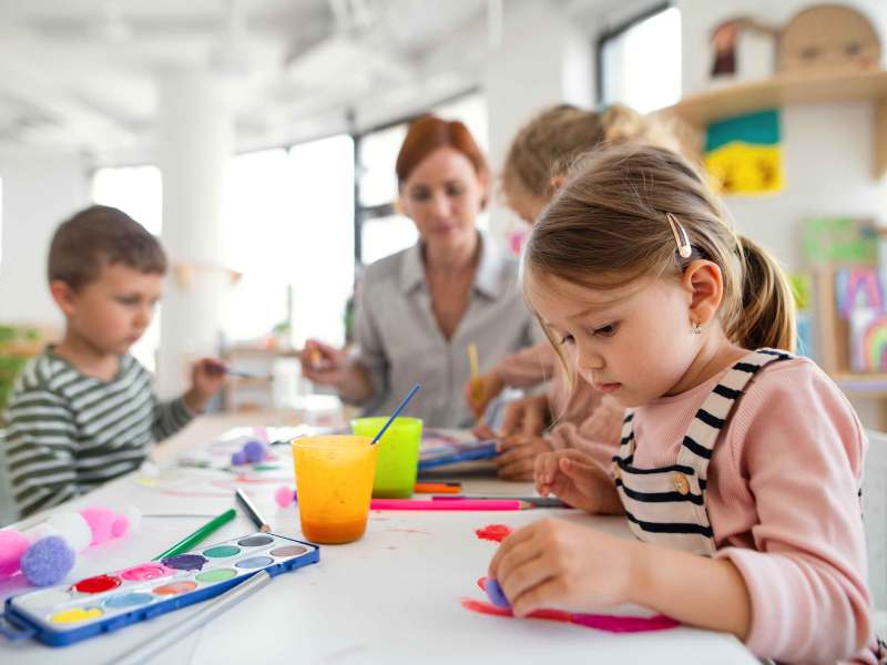 Drei Vorschulkinder sitzen in ihrem Gruppenraum mit einer Erzieherin am Tisch und malen mit Wasserfarben.