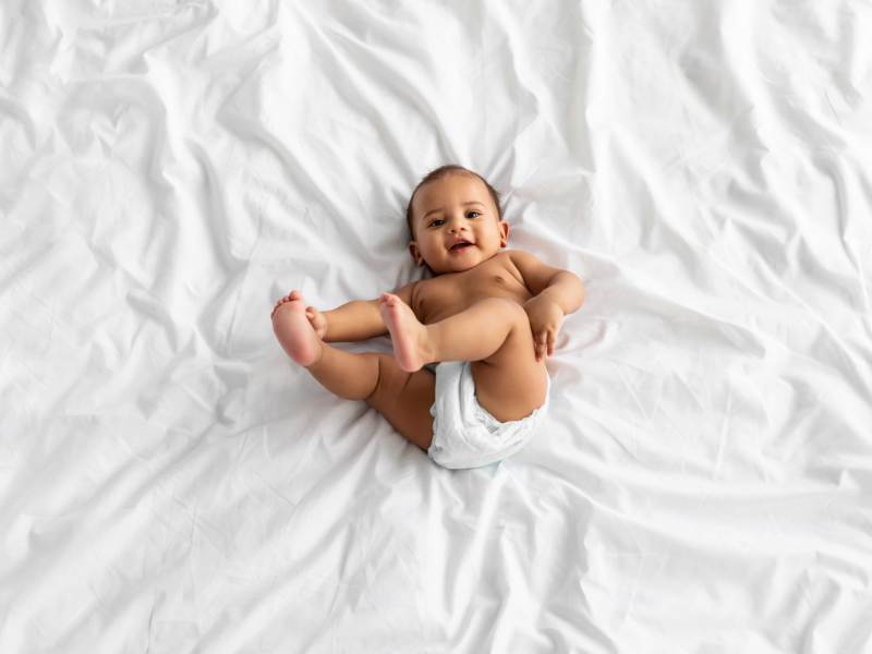 Foto eines Babys von oben, das fröhlich auf einer Decke liegt und mit seinen Füßen spielt.