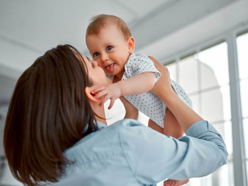 Mutter hebt ihr Baby nach oben und das streckt freudig die Zunge raus.