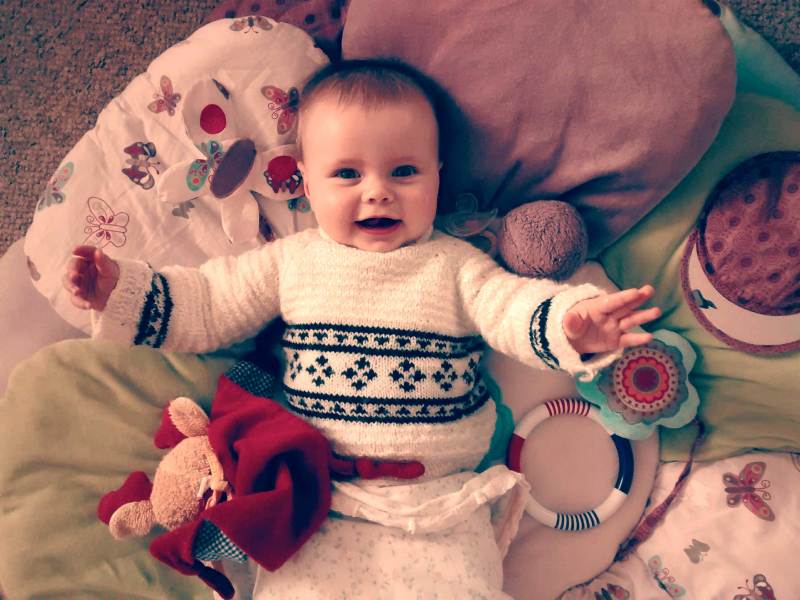 Baby liegt auf einer Decke, umgeben von Spielsachen und lacht in die Kamera.