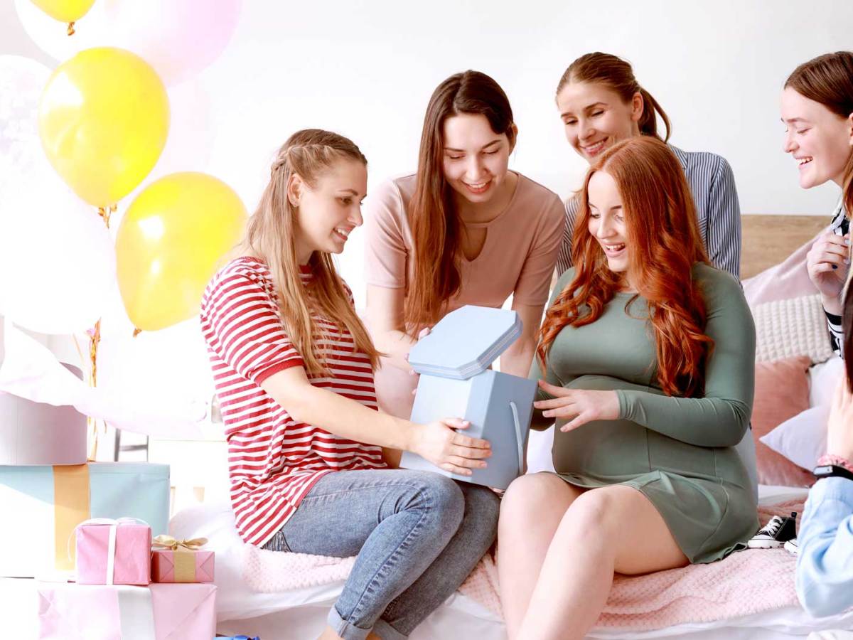 Gruppe Frauen sitzt um ihre schwangere Freundin herum, die gerade in ein Geschenk schaut.