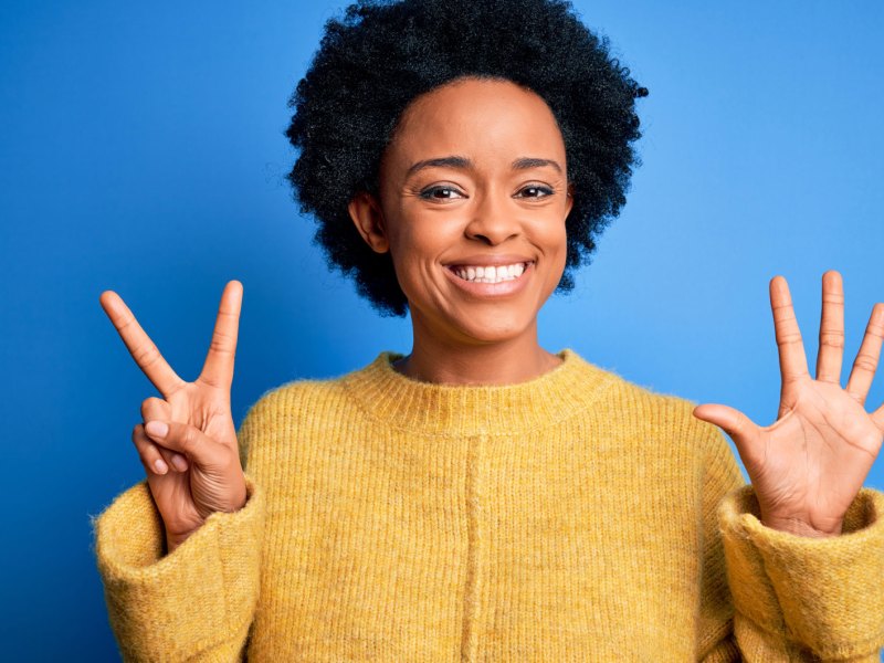 Junge schöne afroamerikanische Frau mit lockigem Haar, trägt einen gelben Freizeitpullover und zeigt mit den Fingern die Nummer sieben, während sie selbstbewusst und glücklich lächelt