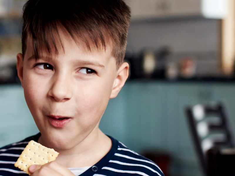 Junge, ca. 10 Jahre alt, schaut verschmitzt zur Seite und isst dabei einen Cracker.