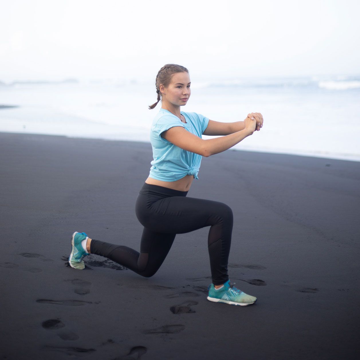Sportliche junge Frau macht auf schwarzem Strand Ausfallschritte. 