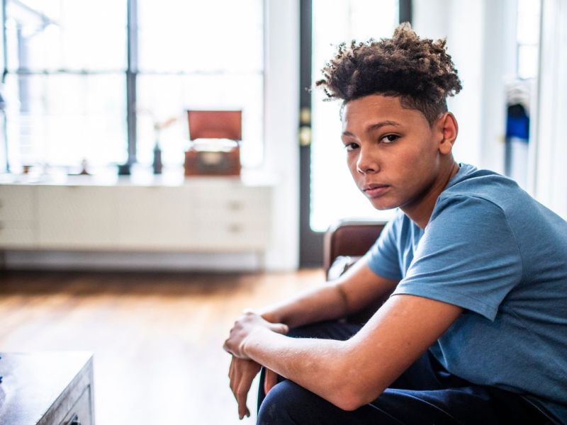 Porträt eines Jungen im Teenageralter, der in einem Wohnzimmer sitzt und ernst in die Kamera schaut.