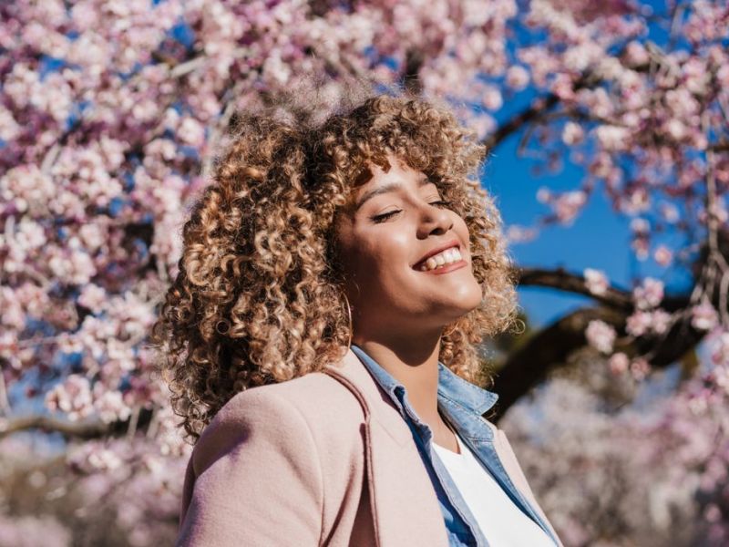 Frau mit Locken steht unter Kirschblüten-Baum und genießt Sonne.