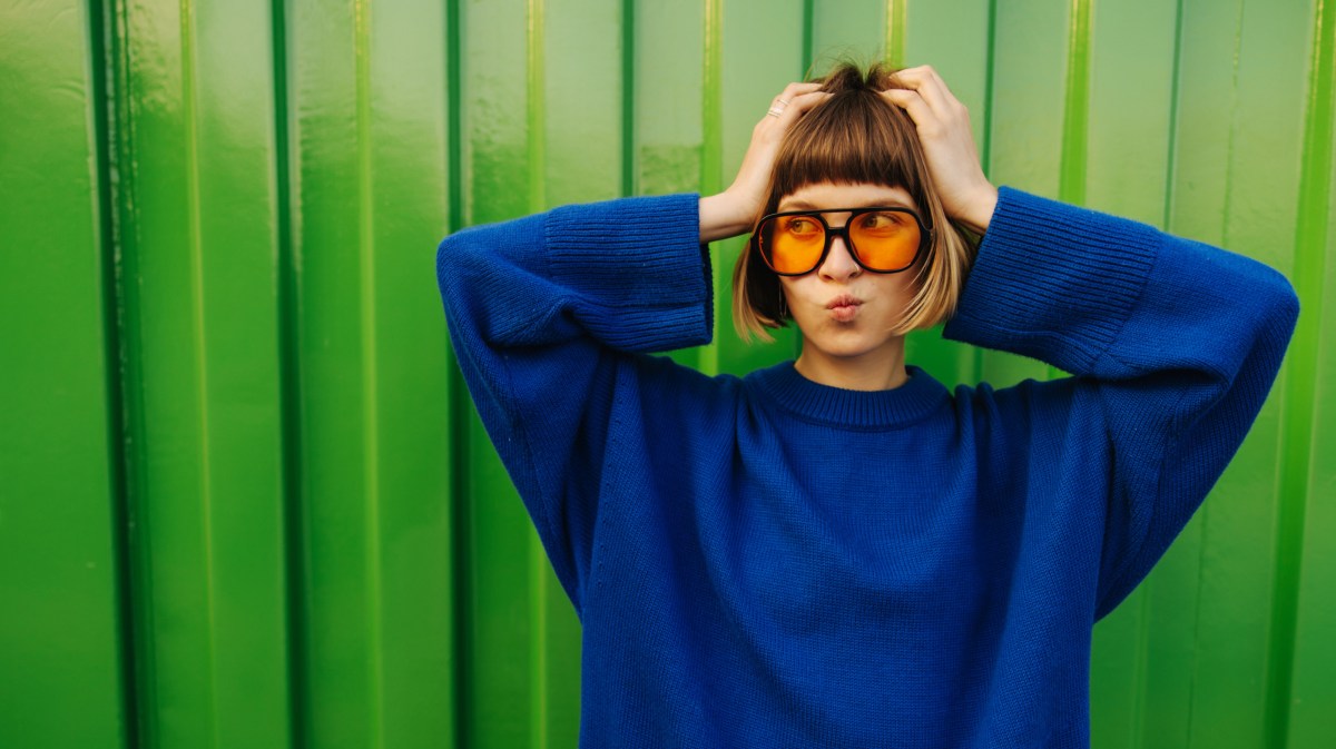 Frua mit Brille und blauem Pullover, die sich in die Haare fasst und vor einer grünen Wand steht
