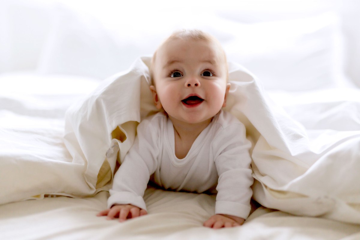 Baby, ca. 7 Monate, liegt unter der Bettdecke auf dem Bett, stützt sich mit den Händen ab und strahlt in die Kamera.