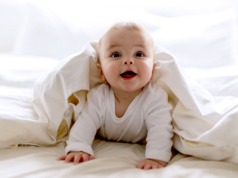 Baby, ca. 7 Monate, liegt unter der Bettdecke auf dem Bett, stützt sich mit den Händen ab und strahlt in die Kamera.