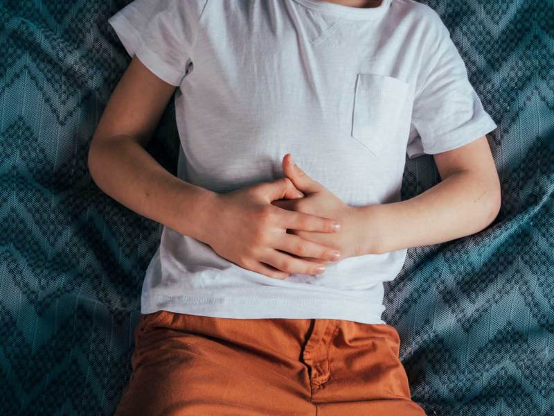 Man sieht den Torso eines Kindes, das auf dem Bett liegt und sich seinen Bauch festhält.