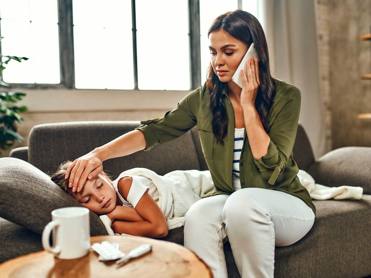 Mutter sitzt auf der Couch neben ihrer kranken Tochter und telefoniert.