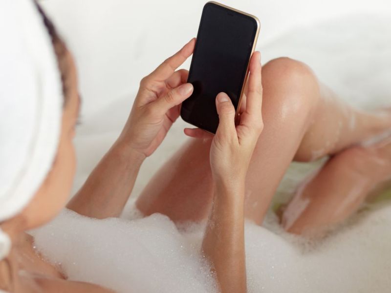 Frau liegt in der Badewanne, mit Smartphone in der Hand