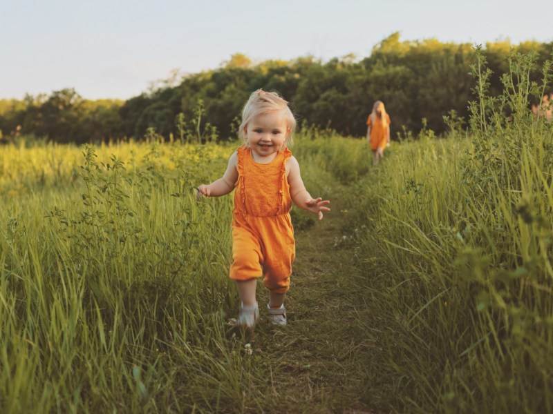 Kleines Mädchen läuft fröhlich über eine Wiese, im Hintergrund steht die Mutter.