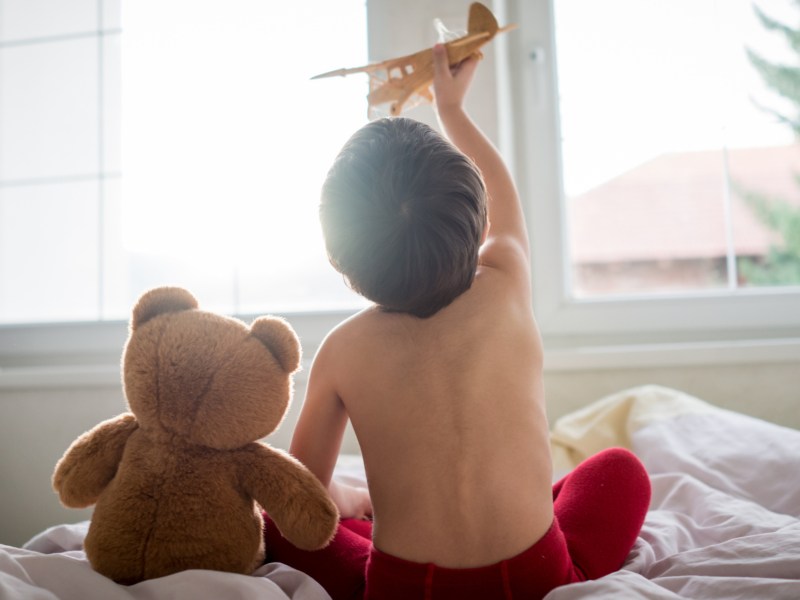 Fröhliches Kind, das mit Holzspielzeugflugzeug und Teddybär im Bett spielt