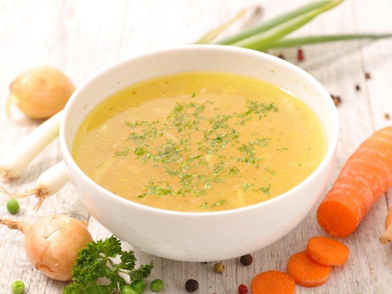 Gemüsebrühe in weißer Suppenschüssel, daneben liegen Karotte, Zwiebel und Lauch.