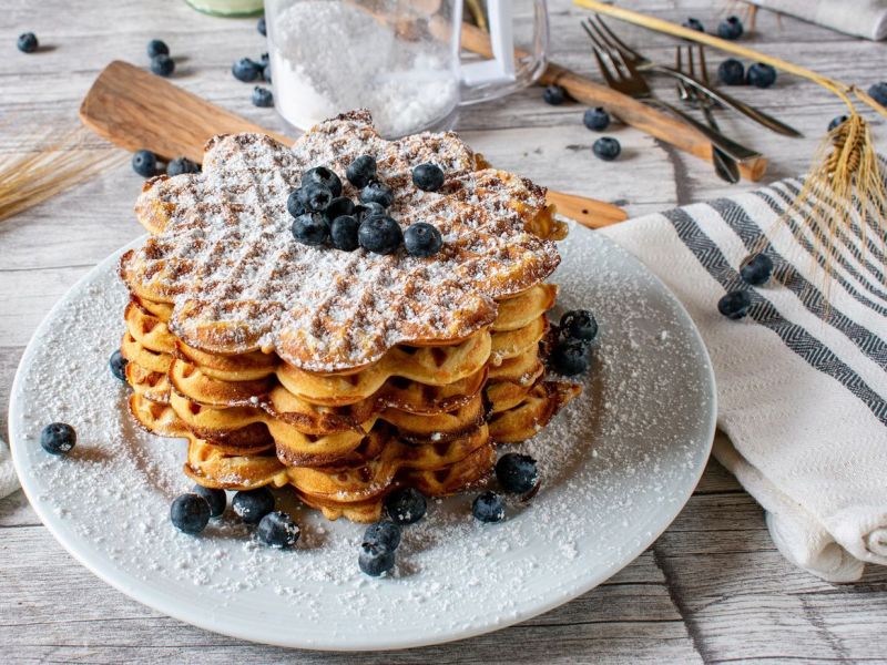 Ein ganzer Haufen Waffeln mit Blaubeeren und Puderzucker auf weißem Teller.