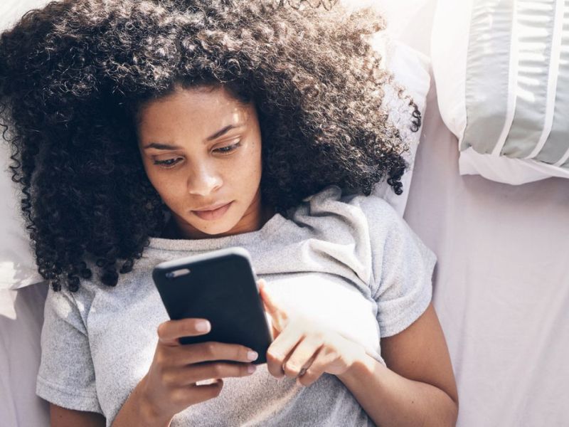 Frau mit dunklen Locken liegt im Bett und schaut auf ihr Handy.