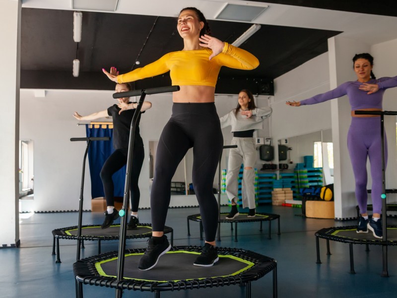 Vier Frauen, die in einem Fitnessstudio auf kleinen Trampolinen springen und dabei Tanzschritte ausführen.