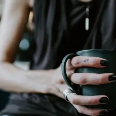 Frau mit kleinem Tattoo am Finger, die eine schwarze Kaffee-Tasse hält