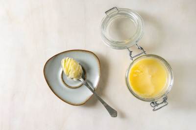Hausgemachtes geschmolzenes Ghee, geklärte Butter in einem offenen Glasgefäß und Löffel auf einer Untertasse auf weißem Marmorhintergrund.