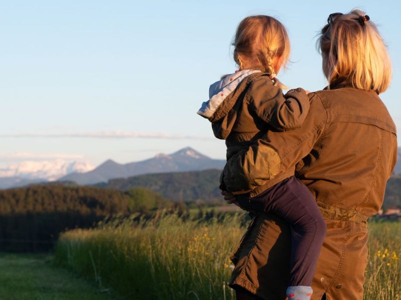 Mutter und Tochter stehen auf einer Wiese und schauen in die Ferne, in der ein Gebirge zu erkennen ist.