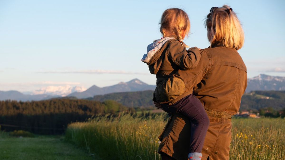 Mutter und Tochter stehen auf einer Wiese und schauen in die Ferne, in der ein Gebirge zu erkennen ist.