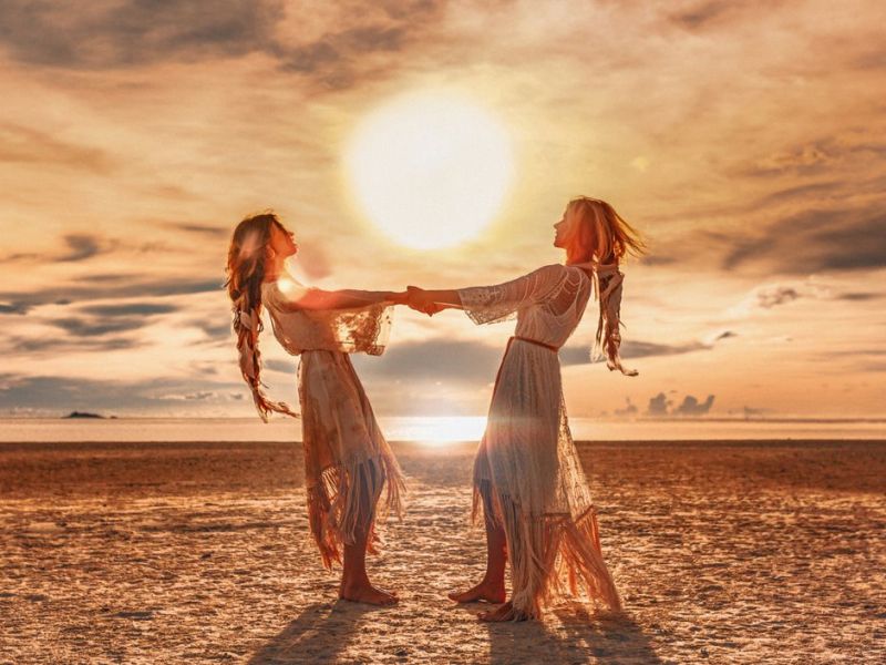Zwei Frauen halten sich am Strand an den Händen, über ihnen die Sonne