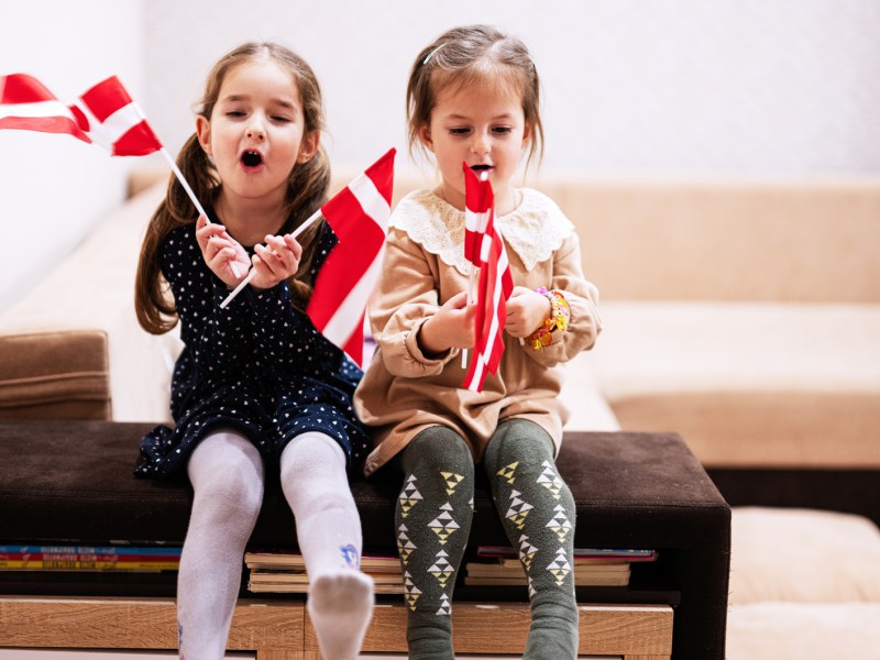 Zwei Mädchen sitzen mit je zwei dänischen Flaggen in den Händen im Wohnzimmer auf einem Hocker.