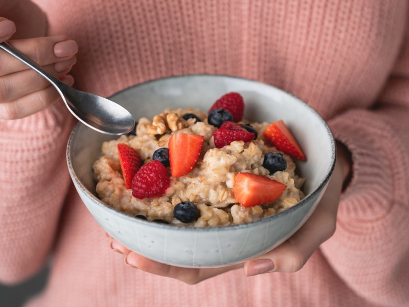 Frau mit rosa Pullover hält Schüssel mit Porridge und Früchten in Hand.