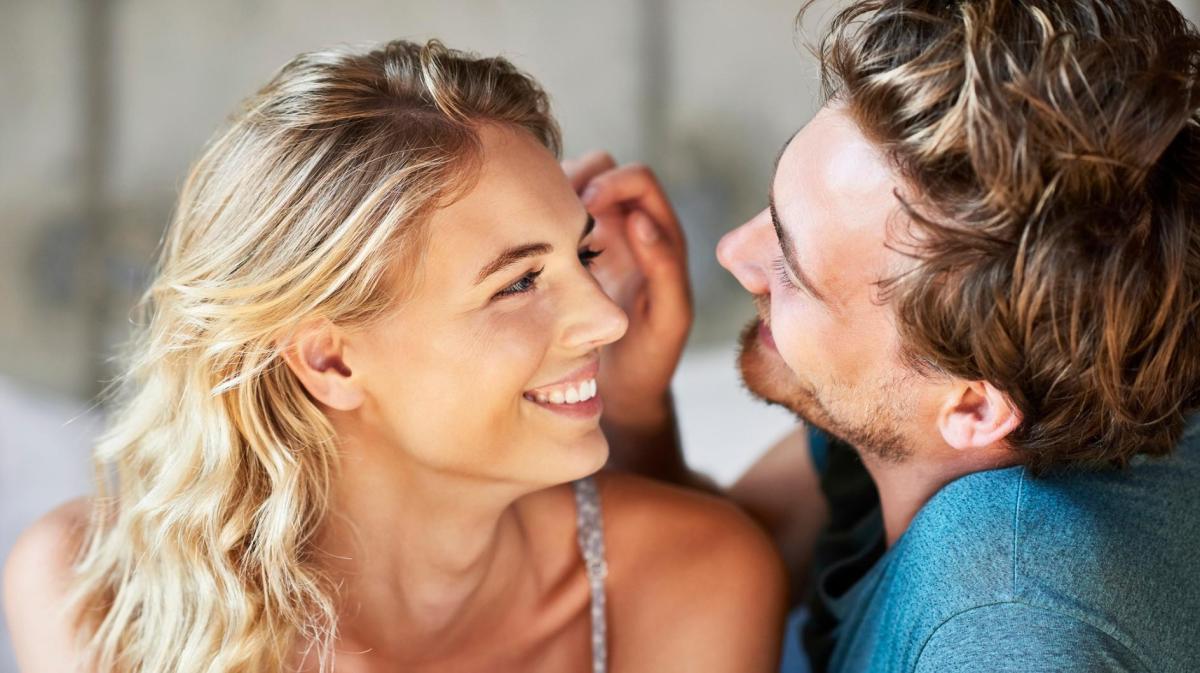 Blonde und Frau und Mann gucken sich verliebt an, während er ihr eine Haarsträhne aus dem Gesicht streicht.