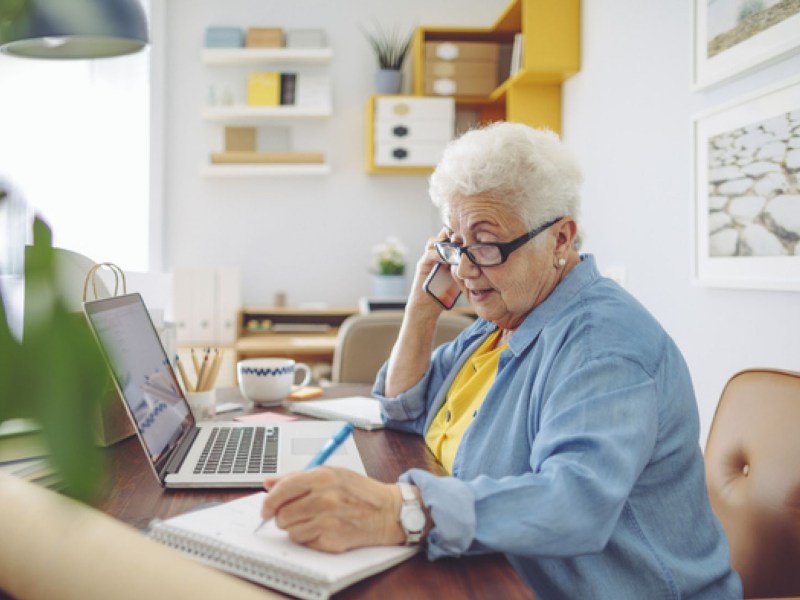 Rentnerinnen sitzt wegen einem Minijob am Laptop und telefoniert.