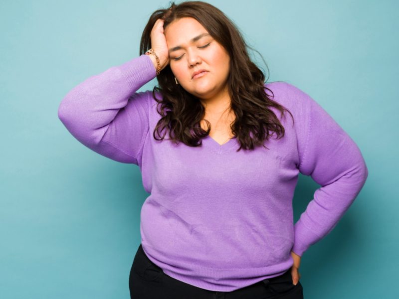 Frau mit lila Oberteil hält sich den Kopf wegen Stress; muss Cortisol senken