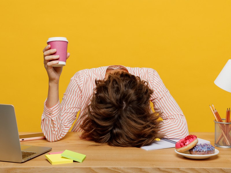 Hohes Cortisol wegen Stress auf der Arbeit; Frau legt Kopf vor gelbem Hintergrund auf die Schreibtischplatte.