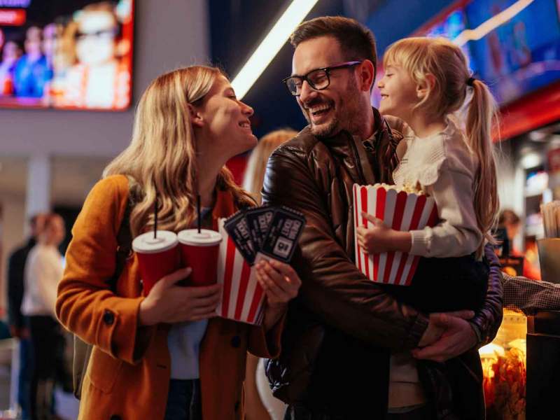 Eltern und Tochter im Kino. Vater trägt seine Tochter, Mutter und Kind halten eine Portion Popcorn in den Armen.
