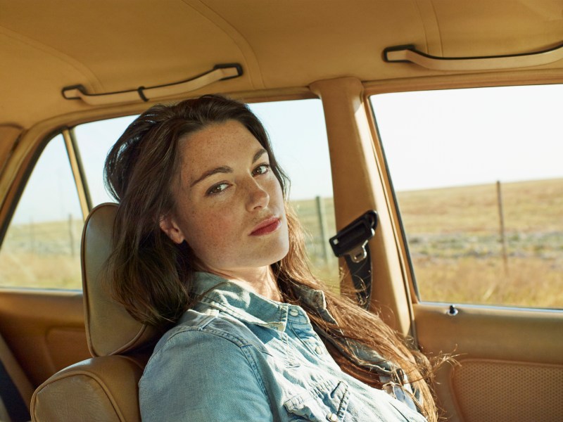 Junge Frau mit langem braunem Haar und hellblauer Jeansbluse sitzt in einem Auto und schaut etwas gelangweilt in die Kamera