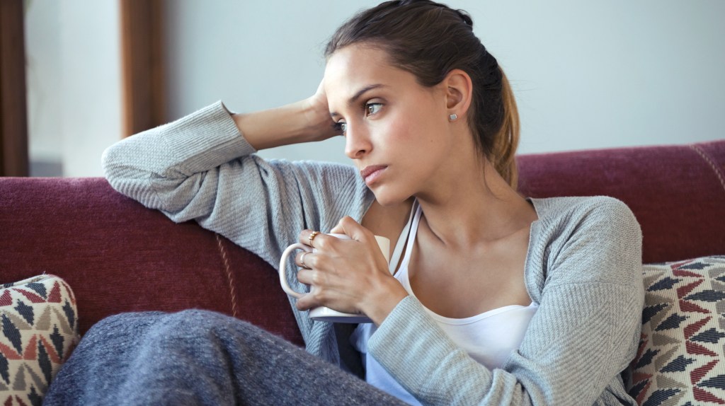 Frau mit hohem Cortisol sitzt auf dem Sofa, Tasse in der Hand.