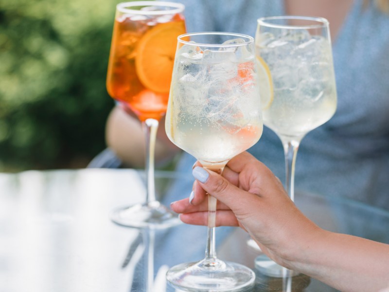 Eine Frau hält einen Limoncello Spritz in der Hand. Im Hintergrund sieht man einen weiteren Limoncello Spritz und einen Aperol Spritz.