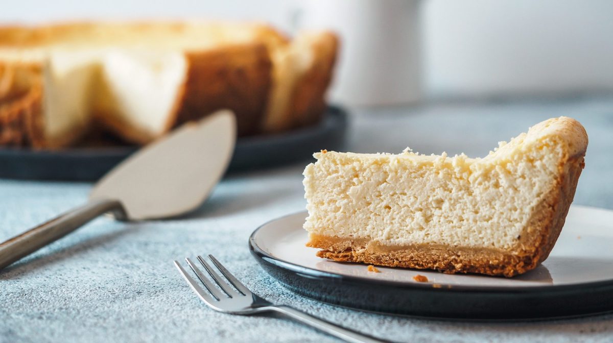 Ein Stück Mango-Vanille-Cheesecake auf einem Teller, daneben eine Gabel, im Hintergrund der angeschnittene Kuchen.
